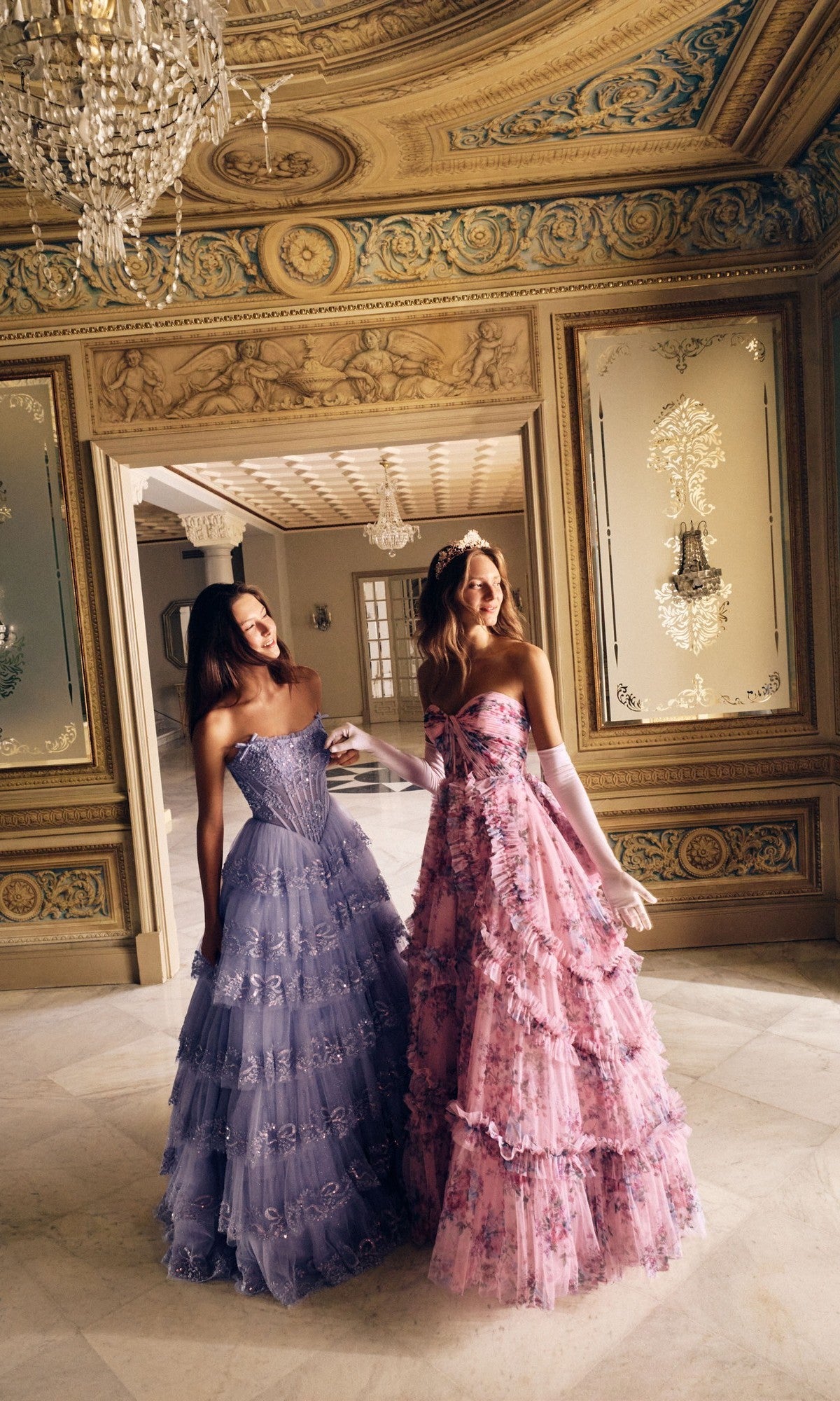 Two women in Alyce 62191 long prom dresses—one with a corset bodice, the other featuring a tulle A-line skirt—smile together in an elegant room adorned with chandeliers, gold trim, and decorative wall panels.